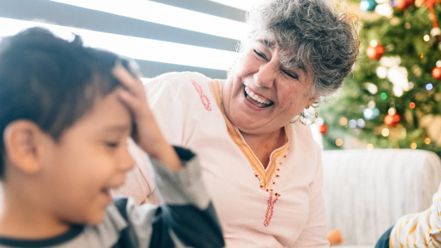 A grandmother and her grandson on a couch. The grandmother's face in crinkled up in laughter as she looks at her young grandson, who is laughing and has his hand against his forehead. A decorated Christmas tree can be seen behind them.